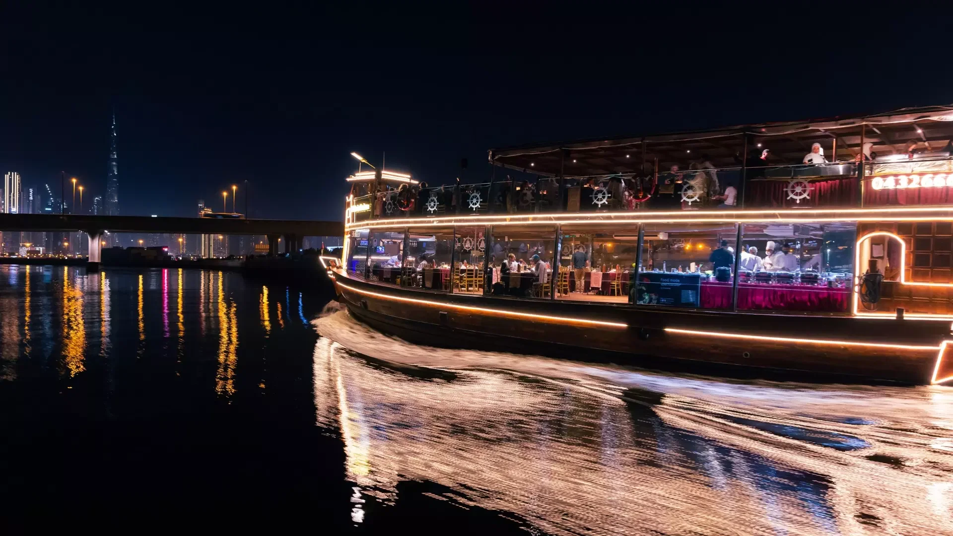 A brightly lit multi-level boat sails along the water at night with city lights visible in the background at Dubai Water Canal Dhow Cruise.