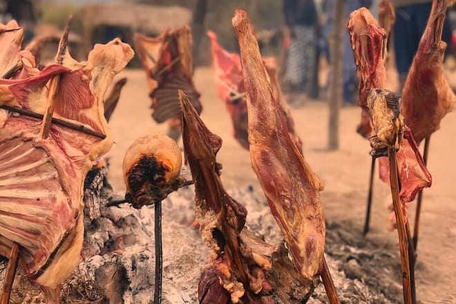 Close-up of large cuts of seasoned meat being slow-grilled on wooden stakes over an open fire in a traditional outdoor setting.