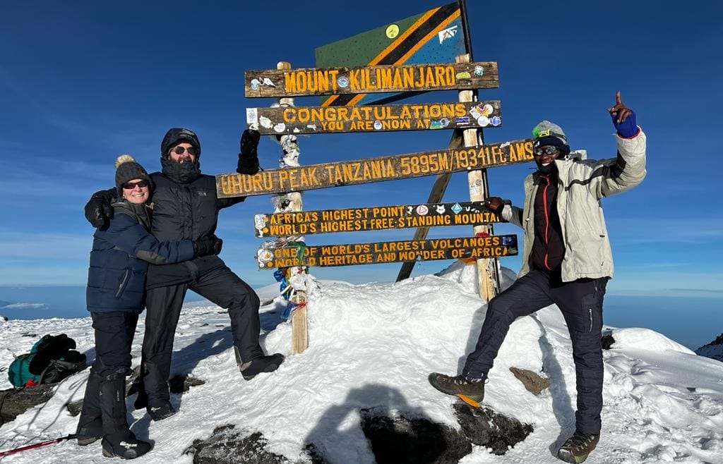 Hikers standing beside the wooden Uhuru Peak sign at the snowy summit of Mount Kilimanjaro under a clear blue sky.