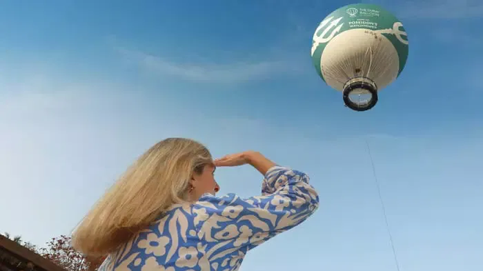 A blond woman in a blue patterned top raises her hand to shield her eyes while looking at a green and white balloon in the blue sky at The Dubai Balloon at Atlantis.