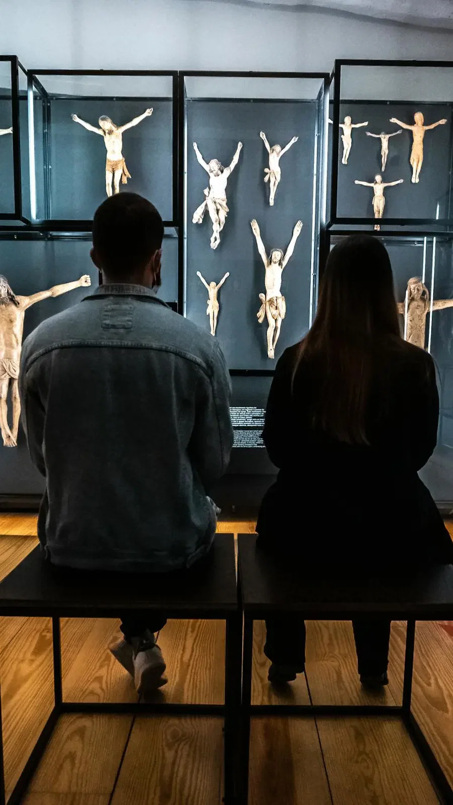 Two people sit on black stools, viewed from behind, looking at a display of various crucifix sculptures in glass cases at Clérigos Tower & Museum Ticket.