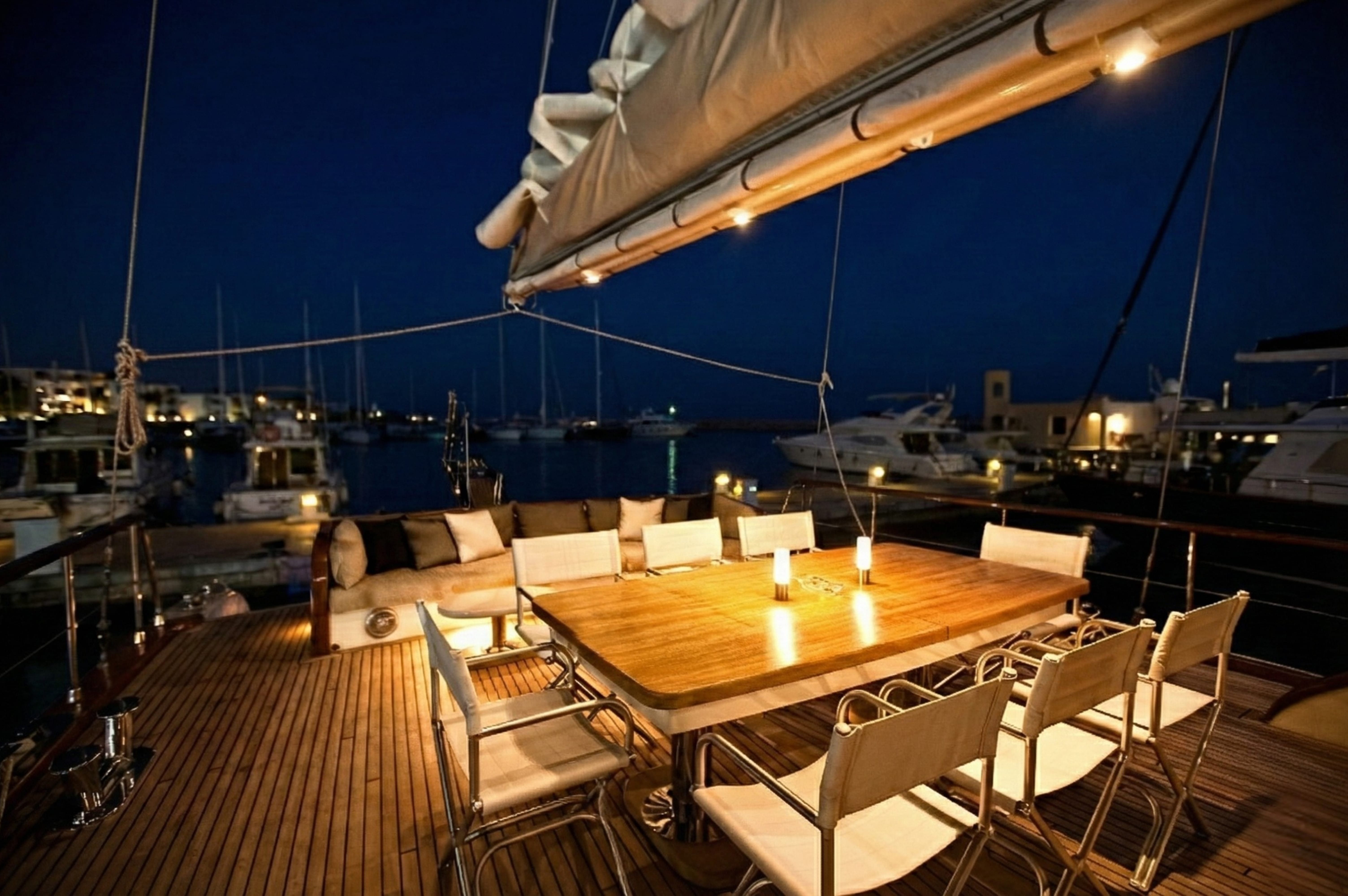 A wooden dining table with chairs and small lamps set up on the open deck of a yacht in Sharm El Sheikh at night.