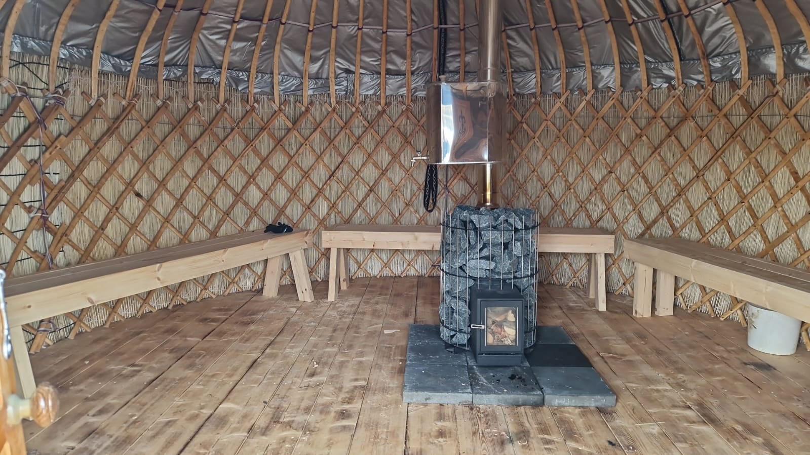 Real interior of the handcrafted yurt sauna with wooden benches, lattice walls and a central wood-burning stone stove.
