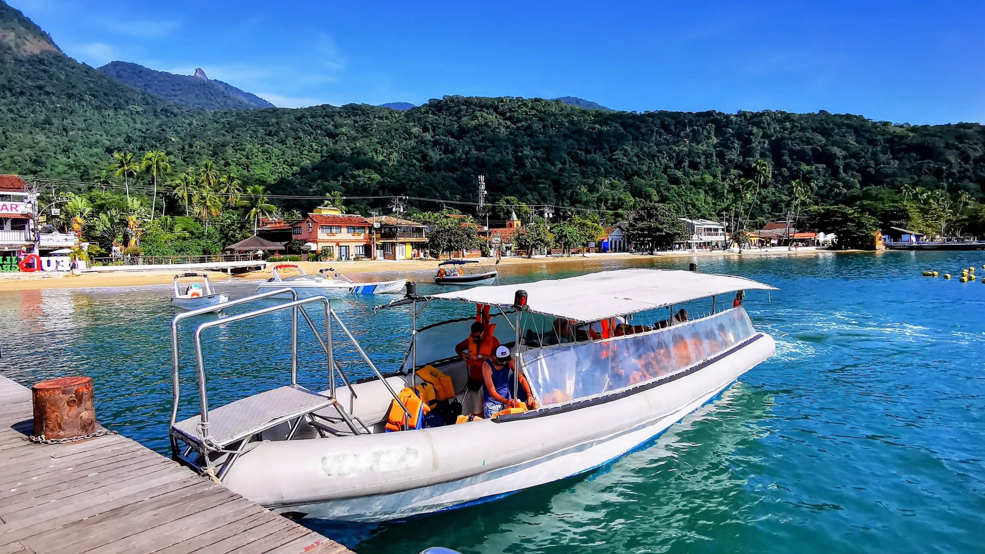 Passageiros embarcando em uma lancha no Píer Turístico na Vila do Abraão, Ilha Grande, com a vila e a Mata Atlântica ao fundo.