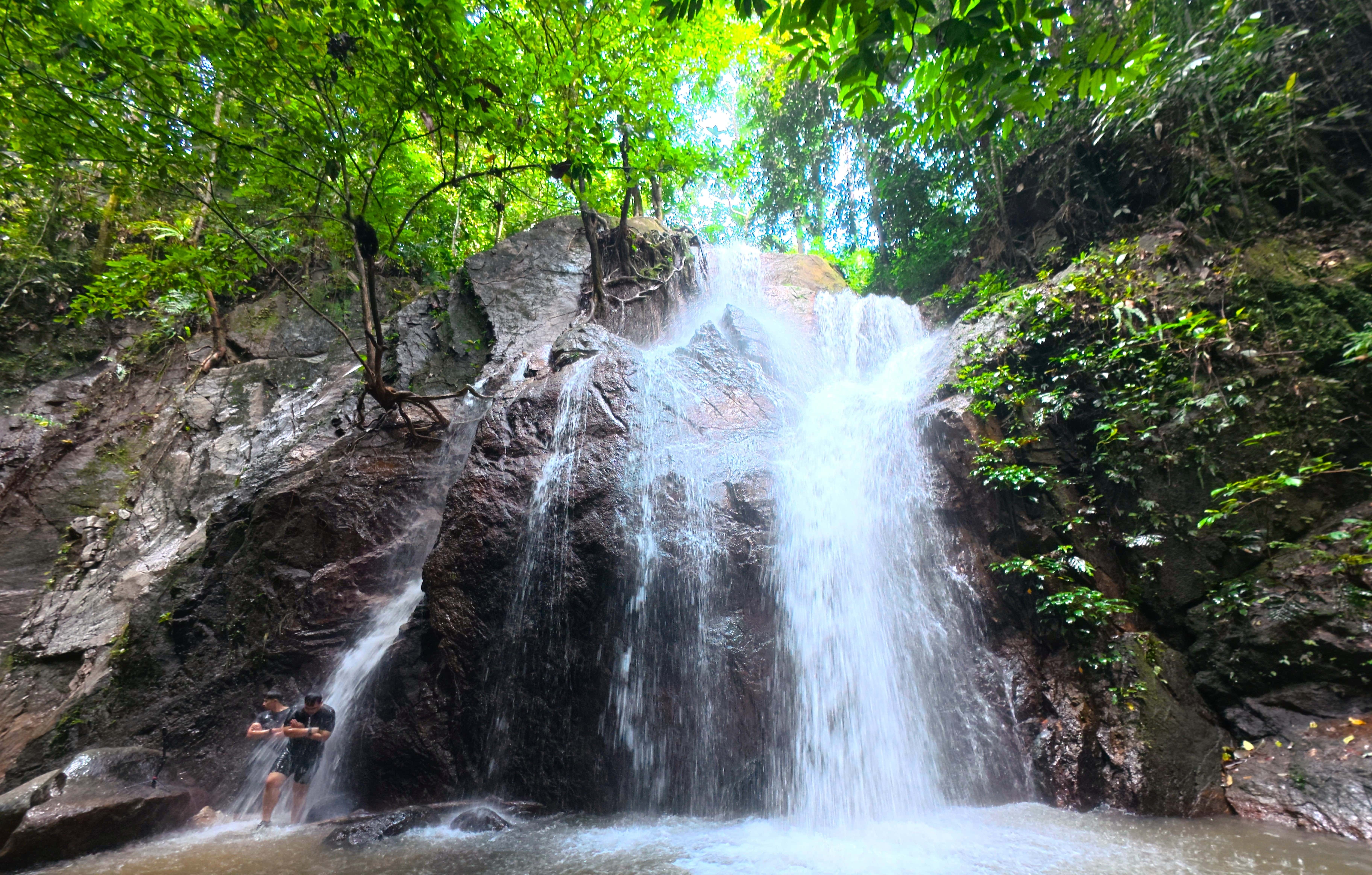 Two hikers enjoying Sungai Pisang Waterfall in Gombak, Selangor, surrounded by lush rainforest and rocky cliffs.