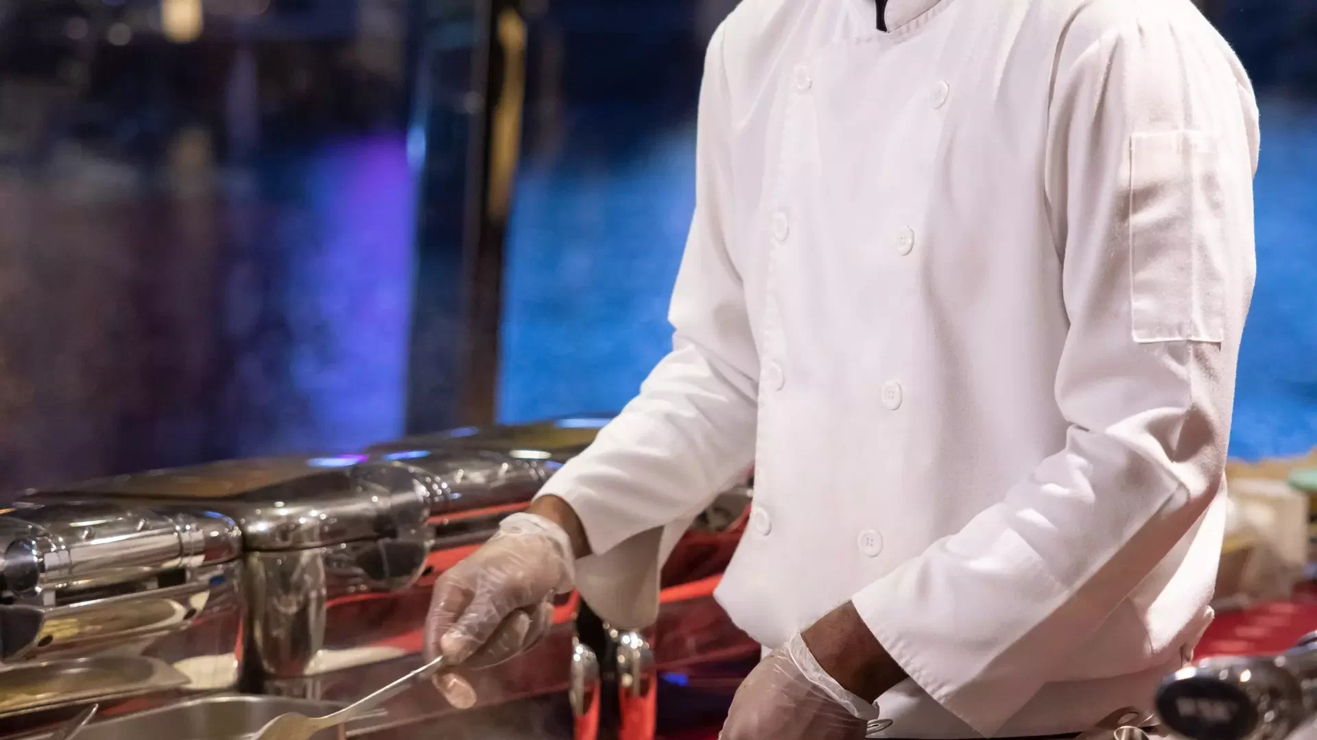 A chef in a white uniform and clear gloves serves food from a buffet line at Dubai Water Canal Dhow Cruise.
