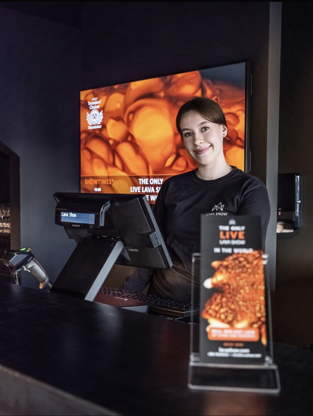 A smiling staff member in a Lava Show t-shirt stands at the ticket and merchandise counter, ready to welcome guests.