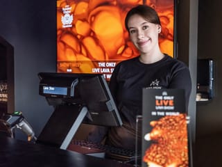 A smiling staff member in a Lava Show t-shirt stands at the ticket and merchandise counter, ready to welcome guests.