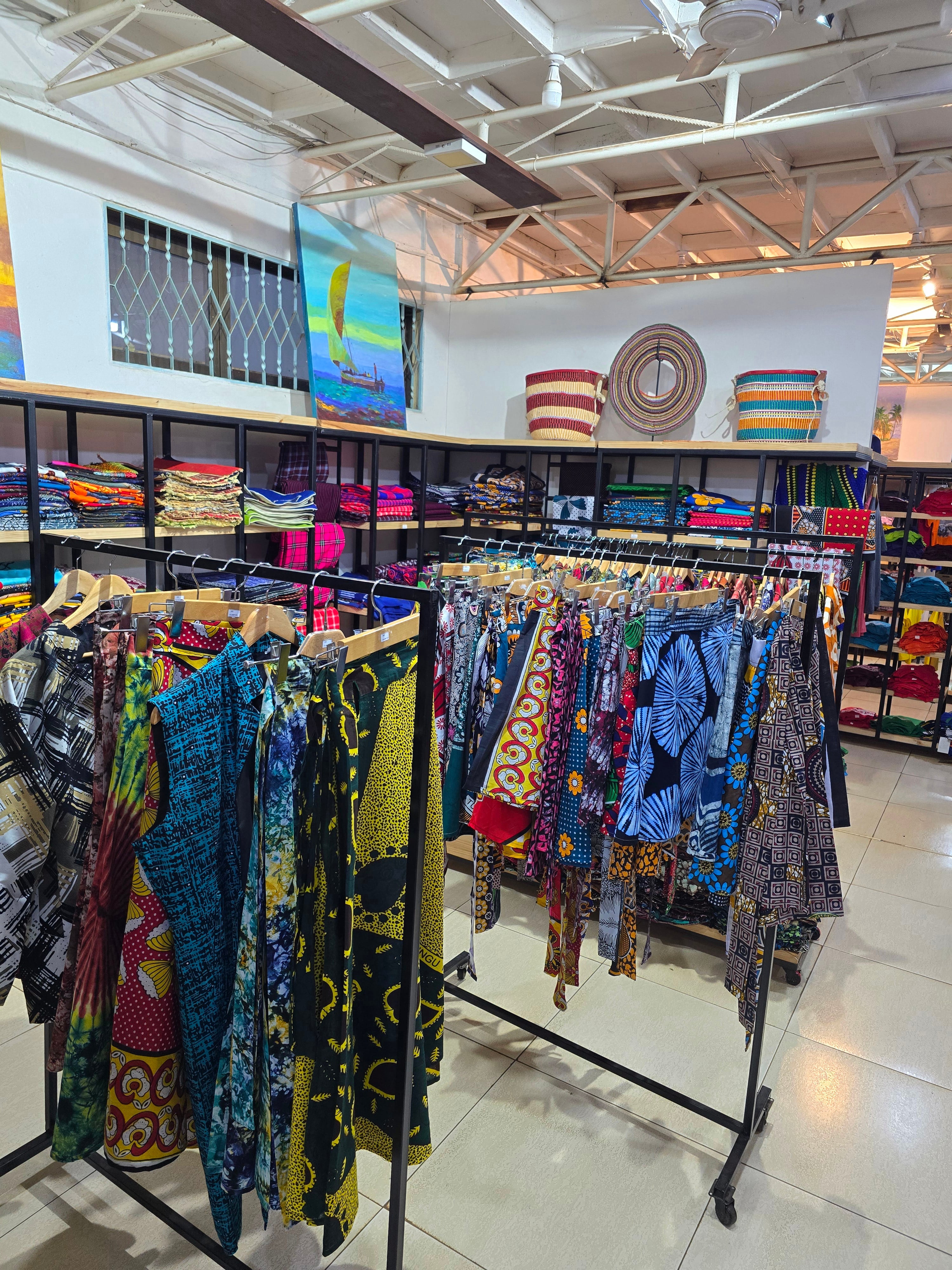 Clothing racks featuring vibrant African print shirts, dresses, and handcrafted textiles in a souvenir shop.