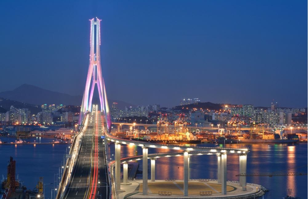 Busan Harbor Bridge illuminated at dusk, with the harbor lights and city skyline glowing along the water.