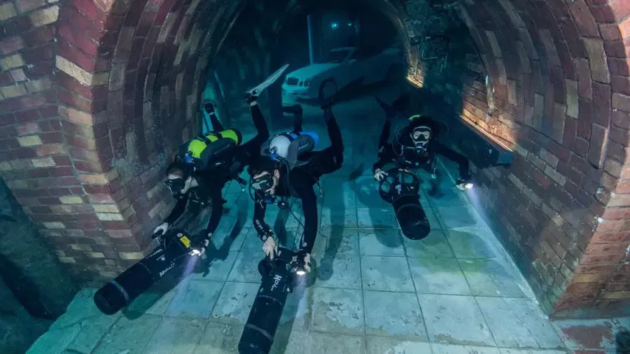 Three divers navigate a submerged brick archway tunnel using underwater propulsion vehicles at Deep Dive Dubai.