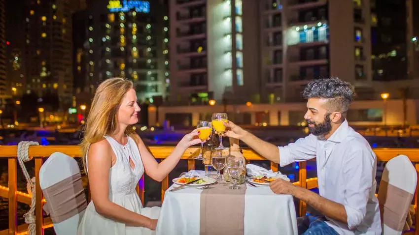 A man and woman sit opposite each other at a candlelit table, smiling and toasting with glasses of orange juice with illuminated city buildings in the background at Alexandra Dhow Cruise in Dubai Marina.