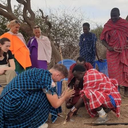A group of tourists and Maasai hosts outdoors watching a guest learn how to make fire using wooden sticks.