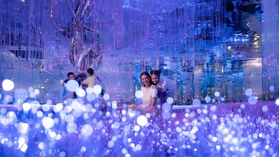 A family sitting in a field of glowing purple and white lights with hanging strands and a mirrored sculpture behind them at AYA Universe Dubai