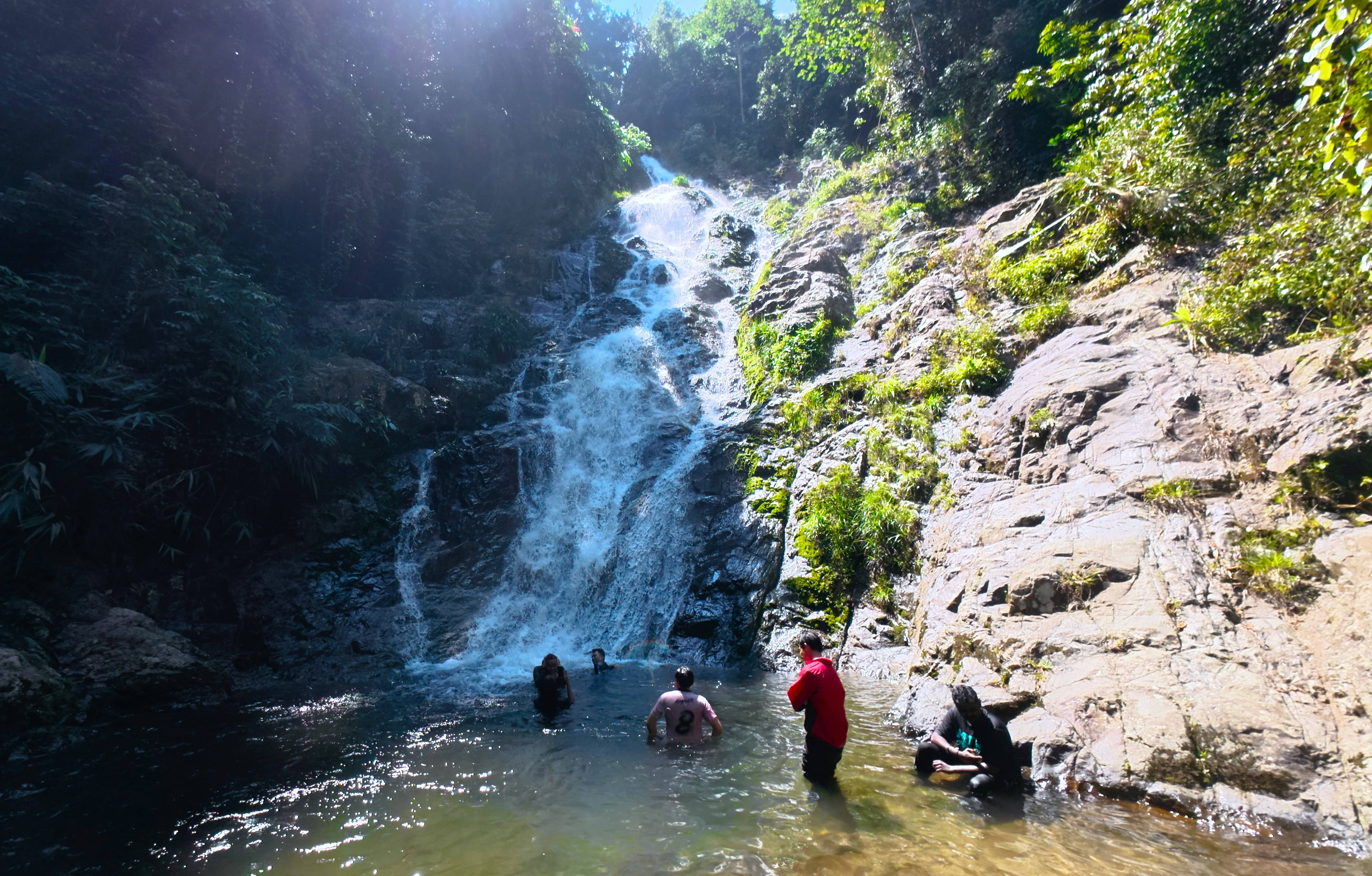 Hikers cool off by swimming in the clear pool at the base of Jeram Perlus waterfall in a lush Malaysian rainforest. 