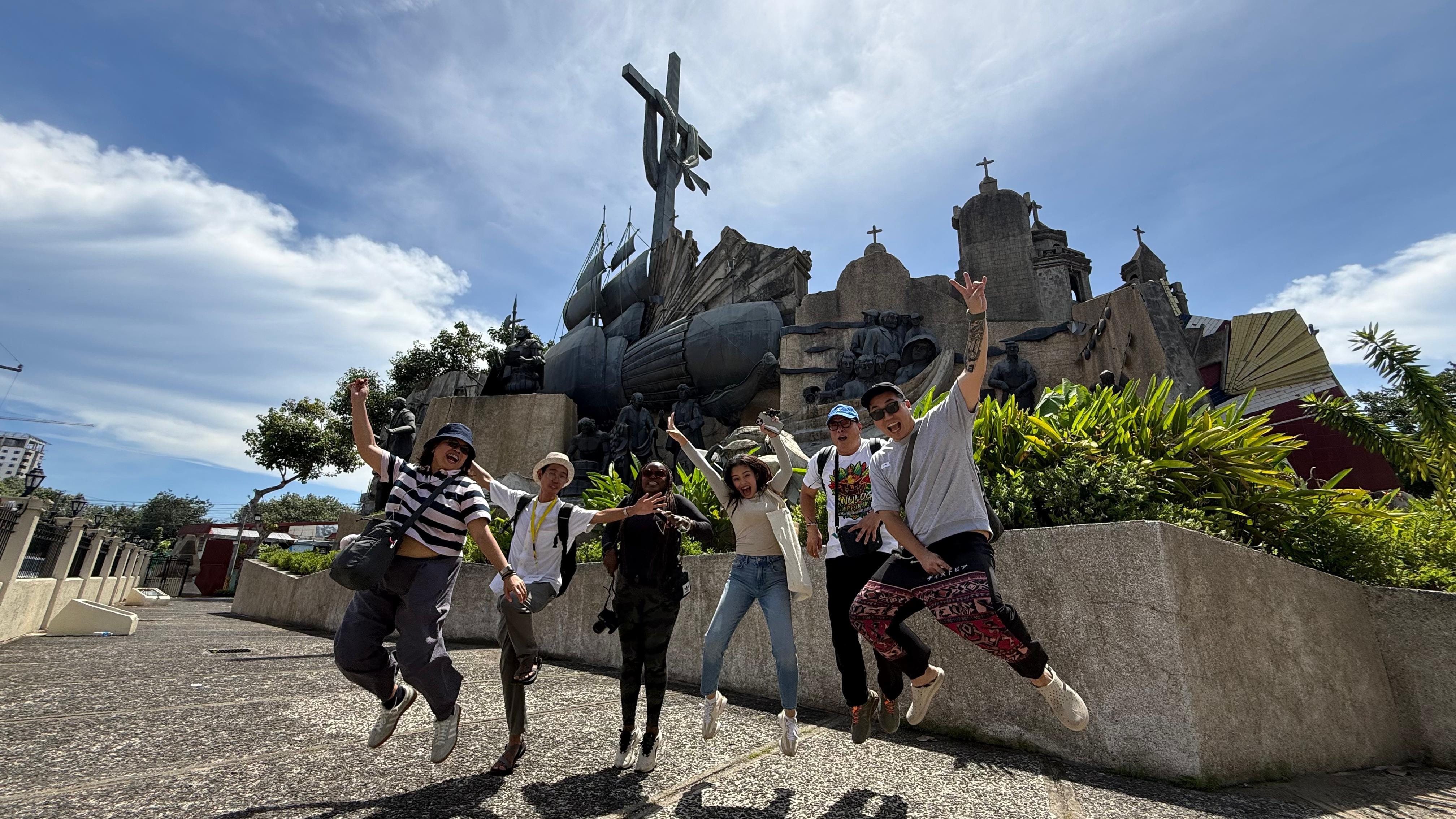 A group of travellers jumps together in front of the Heritage of Cebu Monument during a city tour.