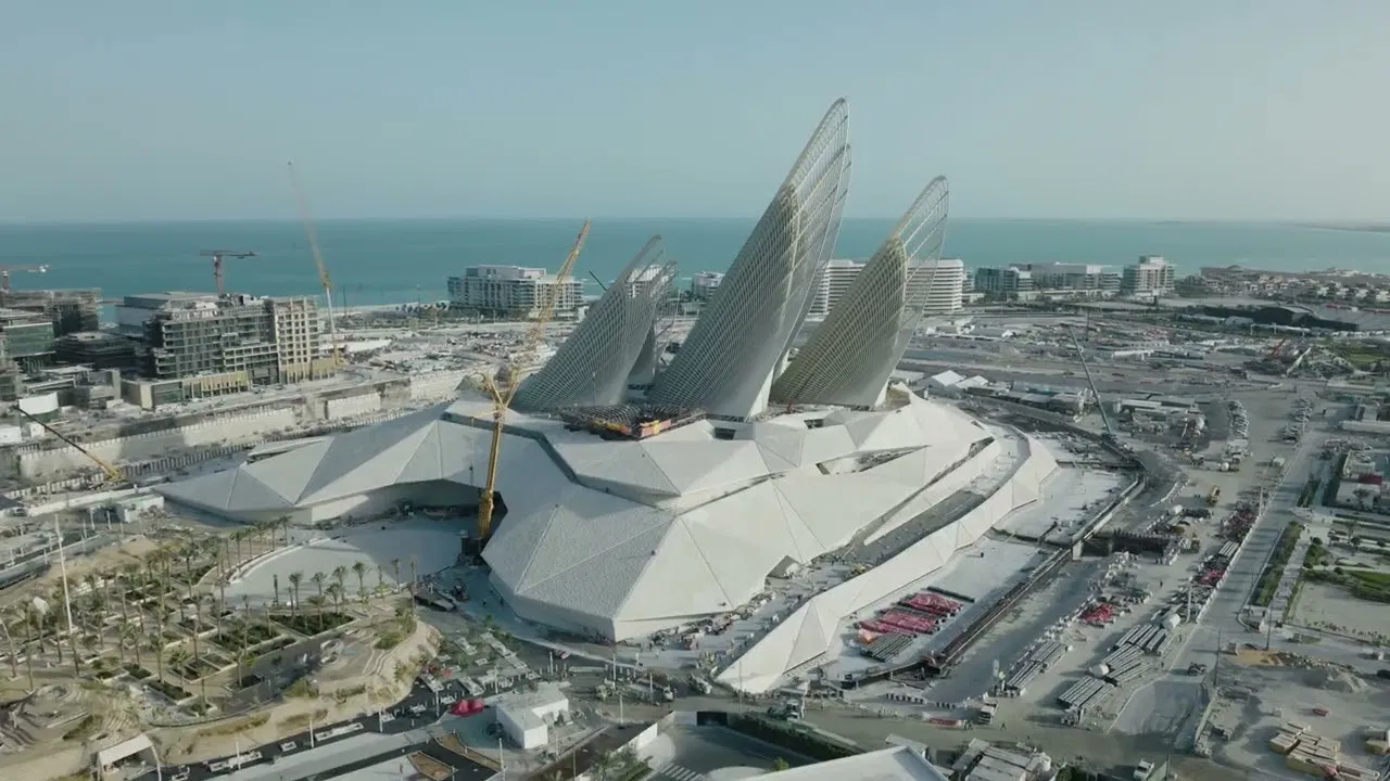 An aerial view shows a large building with wing-shaped structures under construction near the water at Zayed National Museum, Abu Dhabi.