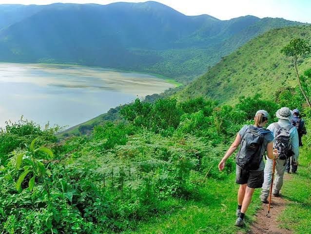 Hikers walking on a narrow dirt path through lush green hills toward a crater lake in the background.
