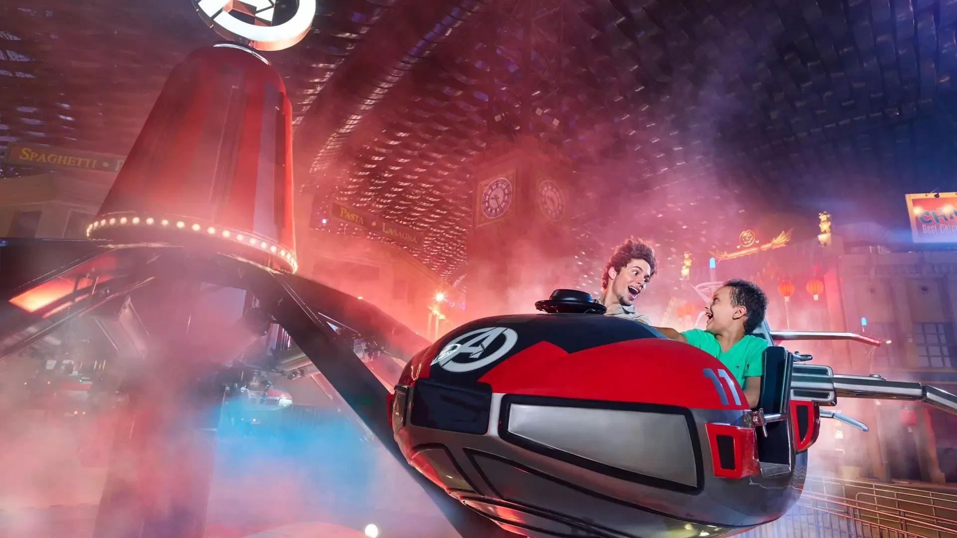 Two smiling visitors ride a spinning red and black ride vehicle beneath a textured ceiling at IMG Worlds of Adventure Theme Park.