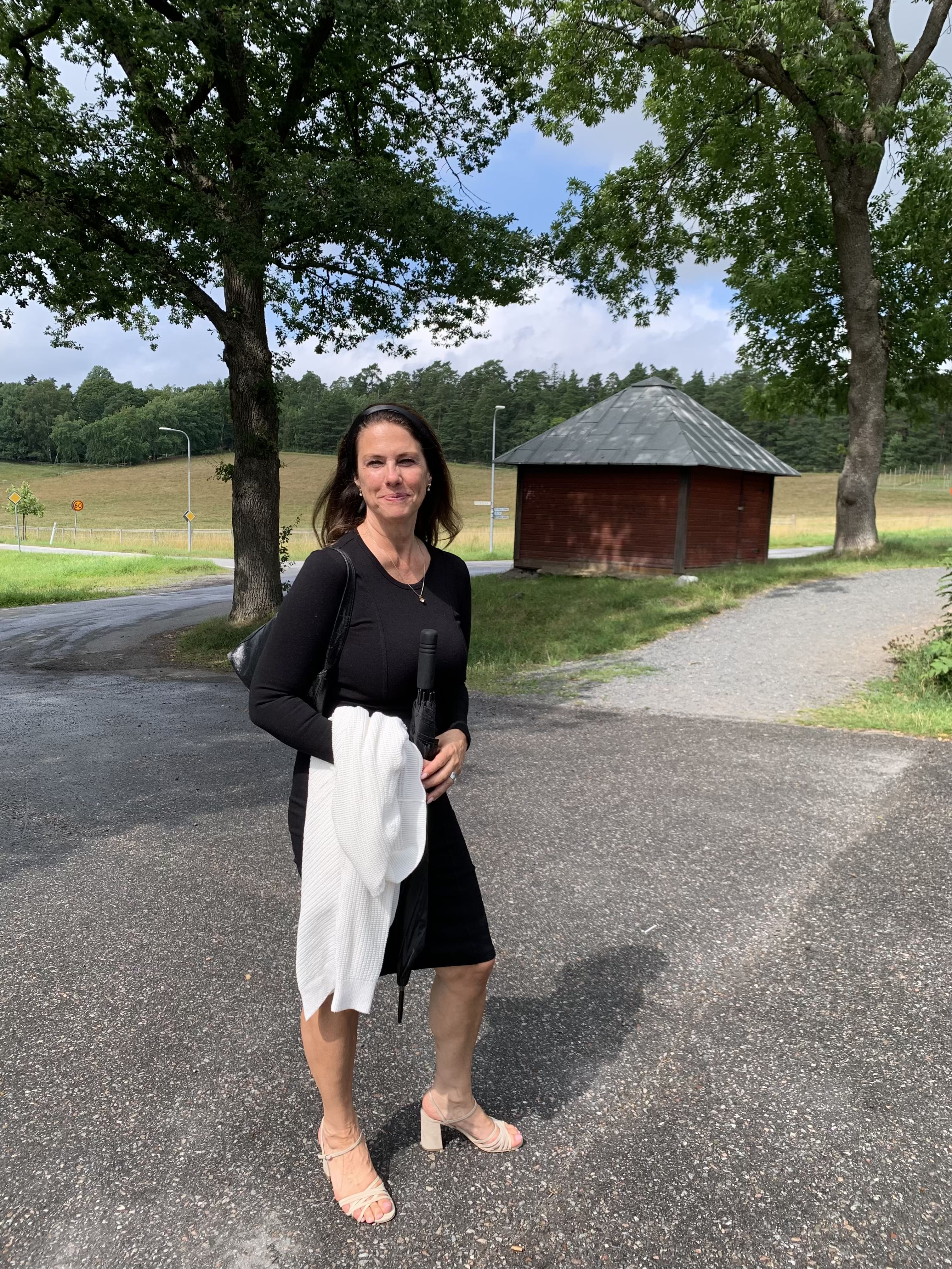 Woman in a black dress and light heels holding a white cardigan, standing on a paved path with fields, trees, and a small red wooden buildin