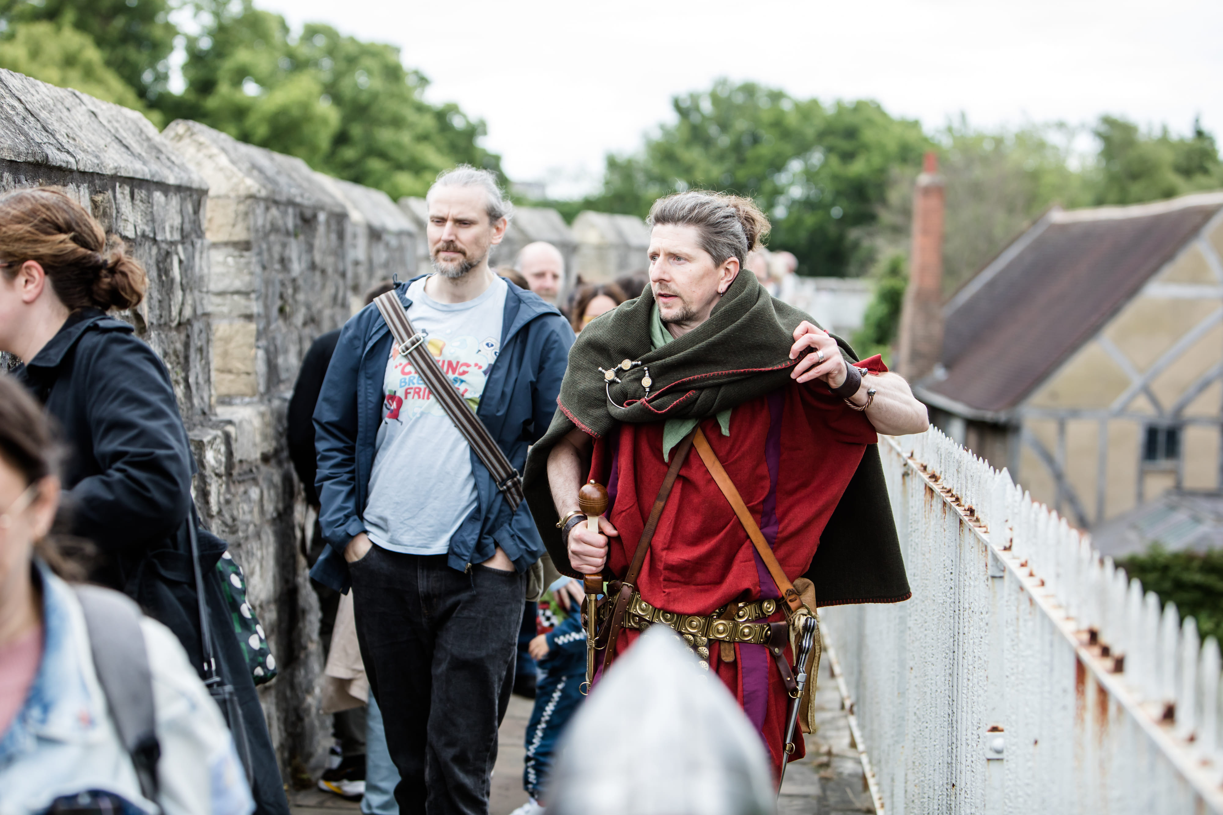 The Original Roman Walk's costumed guide walking along York city walls. There is a medieval house behind him, within the fortress walls.