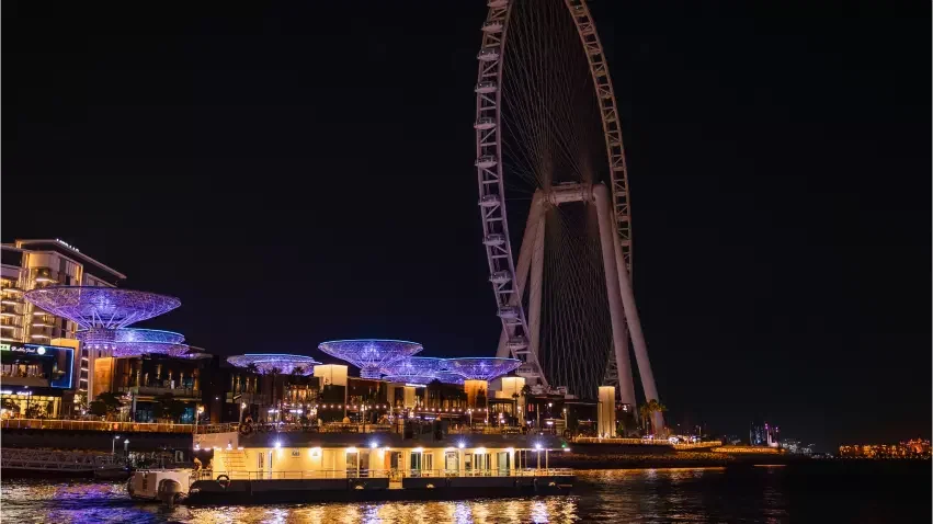 A lighted boat floats on dark water in front of a sprawling city skyline and a massive observation wheel at Alexandra Sea Lounge.