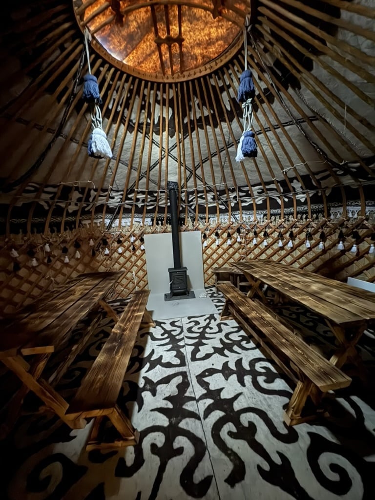 Interior of a traditional dining yurt with two wooden tables, benches and a central wood-burning stove.