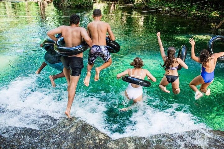 A group of people jump into a clear blue lagoon wearing swimwear and inflatable tubes, splashing into the water surrounded by lush green veg