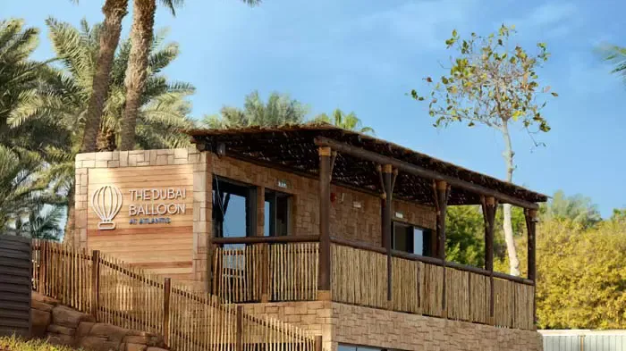 A wooden building with a thatched roof and a stone and bamboo porch is nestled amongst tall palm trees and lush greenery at The Dubai Balloon at Atlantis.