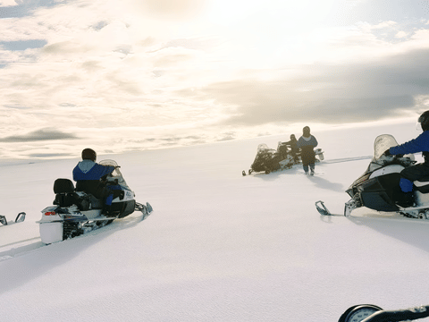 Several snowmobilers in blue suits riding across the wide flat expanse of Langjokull glacier toward a bright overcast horizon, with a guide