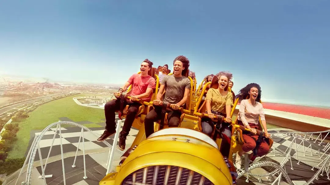 Riders in an open yellow roller coaster train screaming as it crests high above green parkland beside the red roof at Ferrari World Abu Dhabi