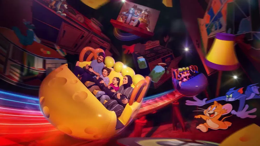 A family rides a yellow cheese-shaped coaster vehicle along a track surrounded by colorful cartoon illustrations at Warner Bros. World Abu Dhabi Theme Park.