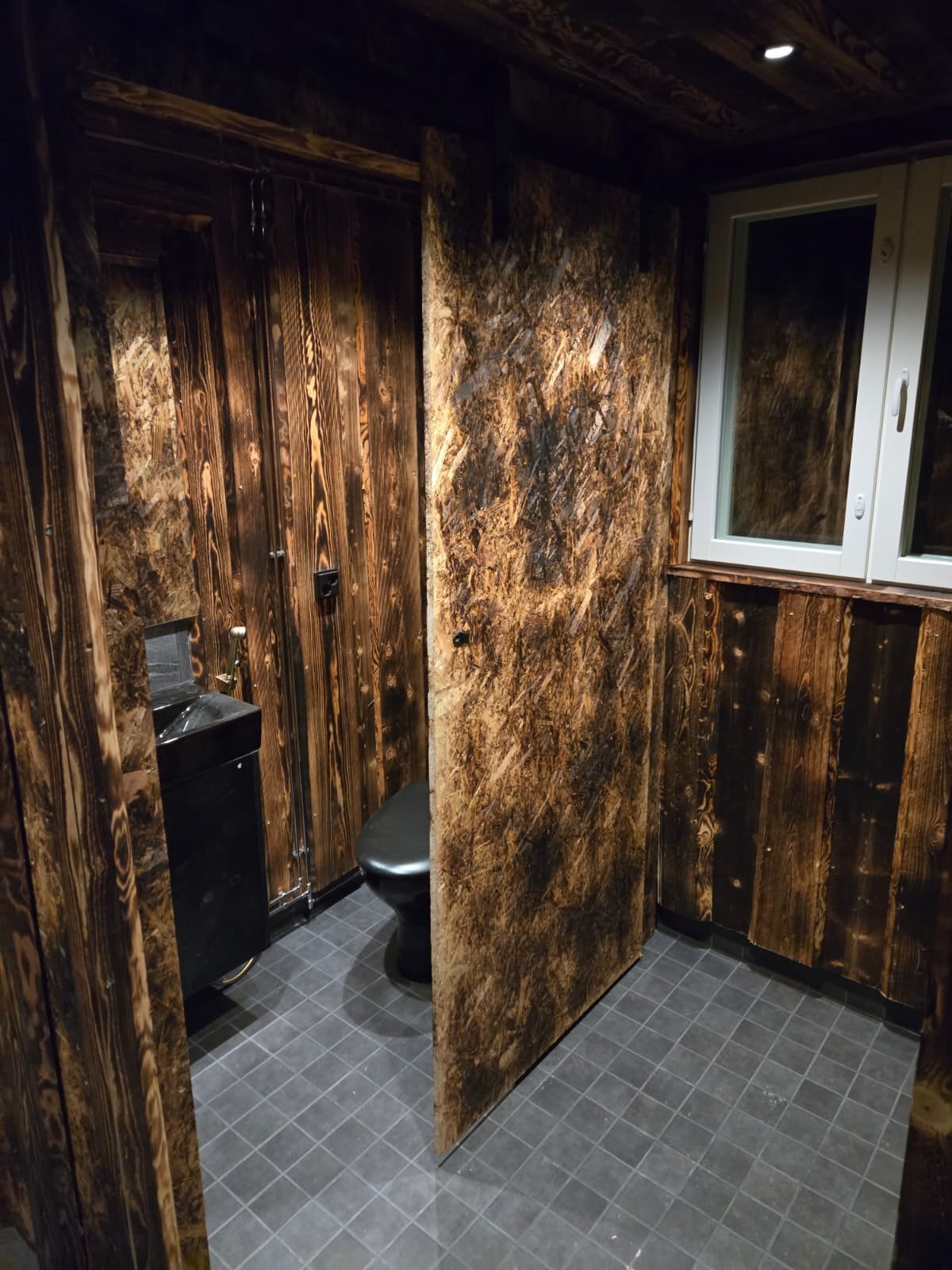 Private WC with dark Finnish timber walls, black fixtures and a window overlooking the forest