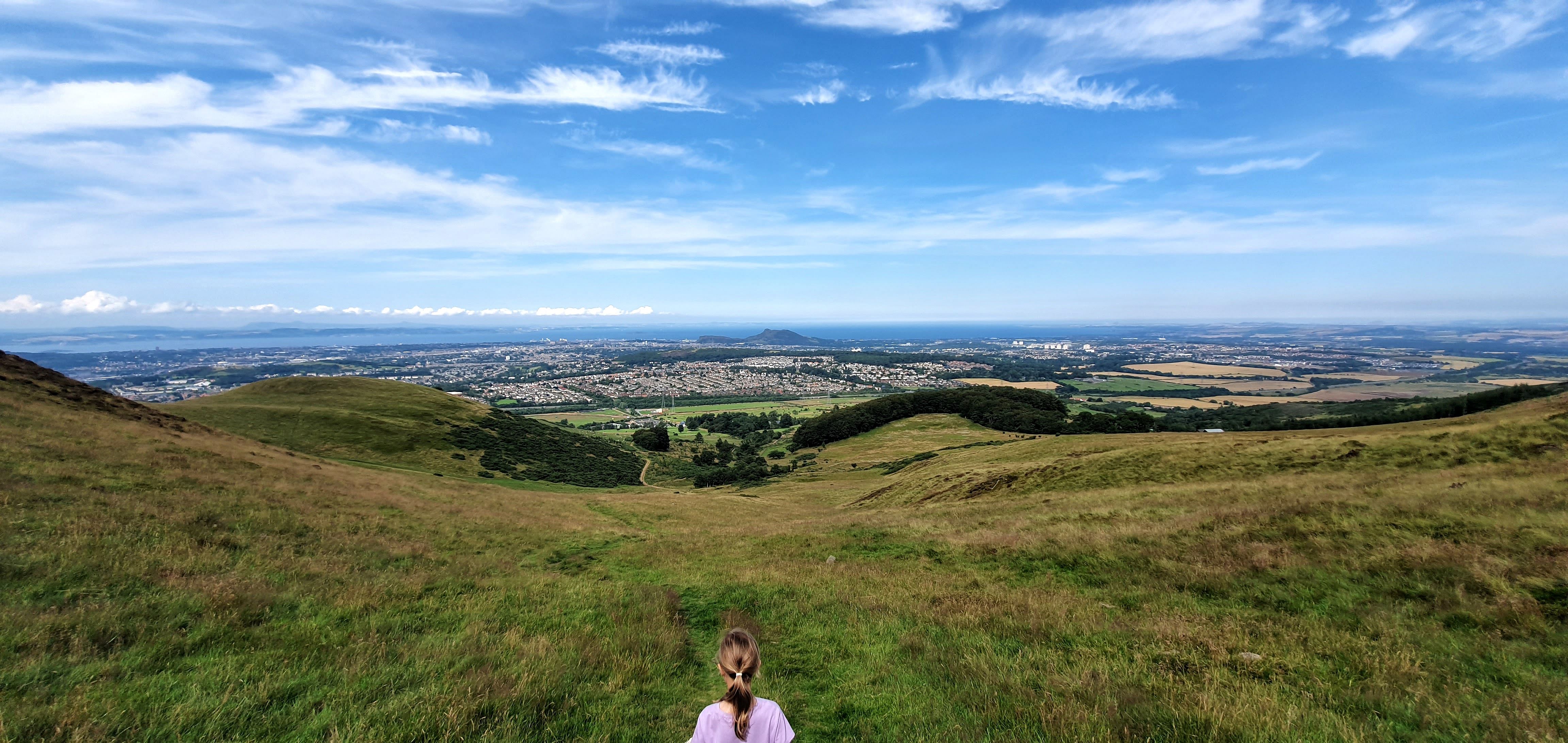 Girl looking at the city of Edinburgh from a hilltop, on a Summers day. Sky is blue and the sea can be seen on the horizon.