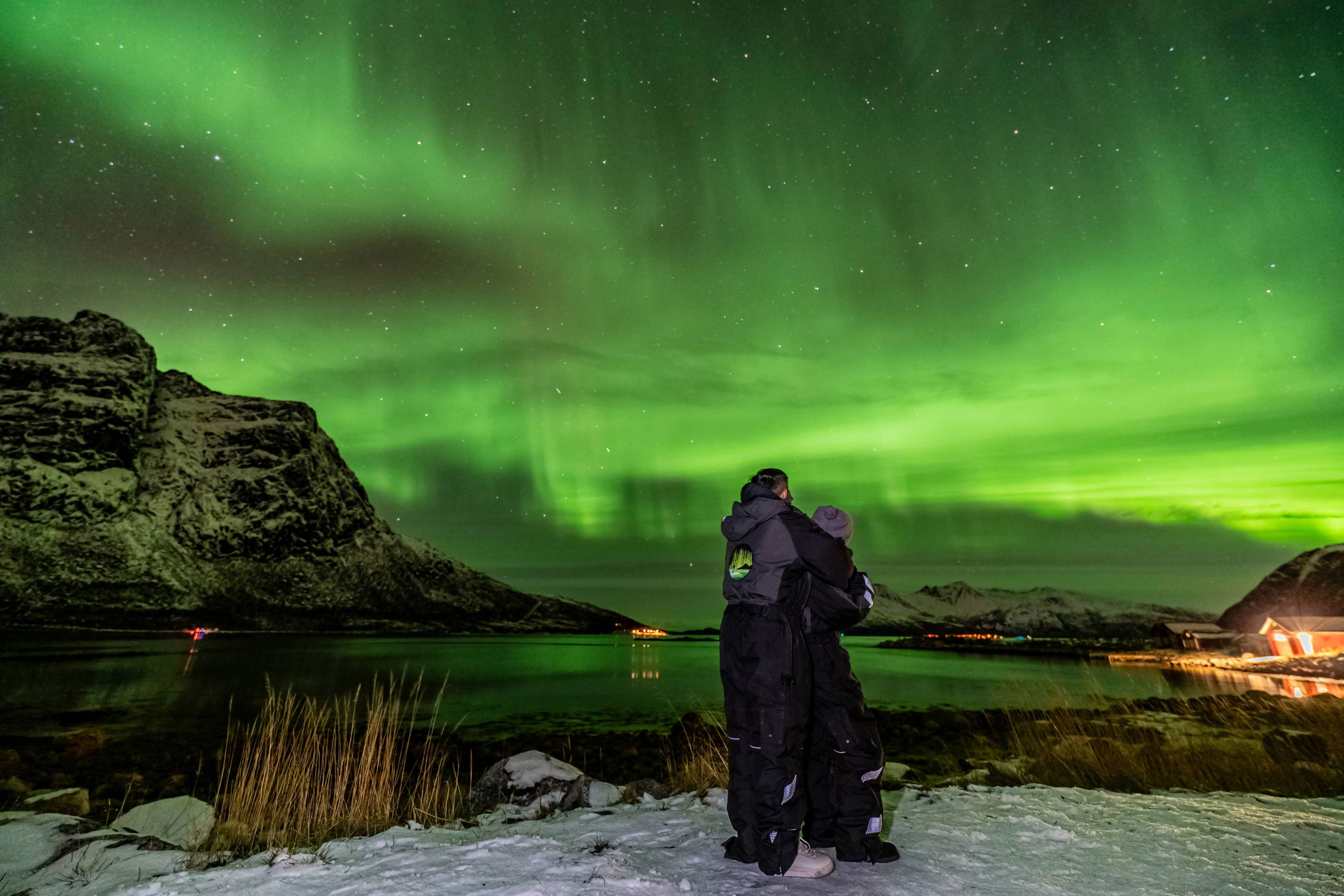 Couple in thermal suits embracing by a rocky Arctic fjord beneath a bright green Northern Lights display on a private Tromsø tour.