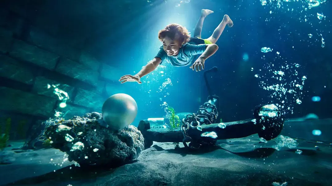 A young boy dives underwater towards a large glowing pearl resting near an anchor at Yas Waterworld, Abu Dhabi.