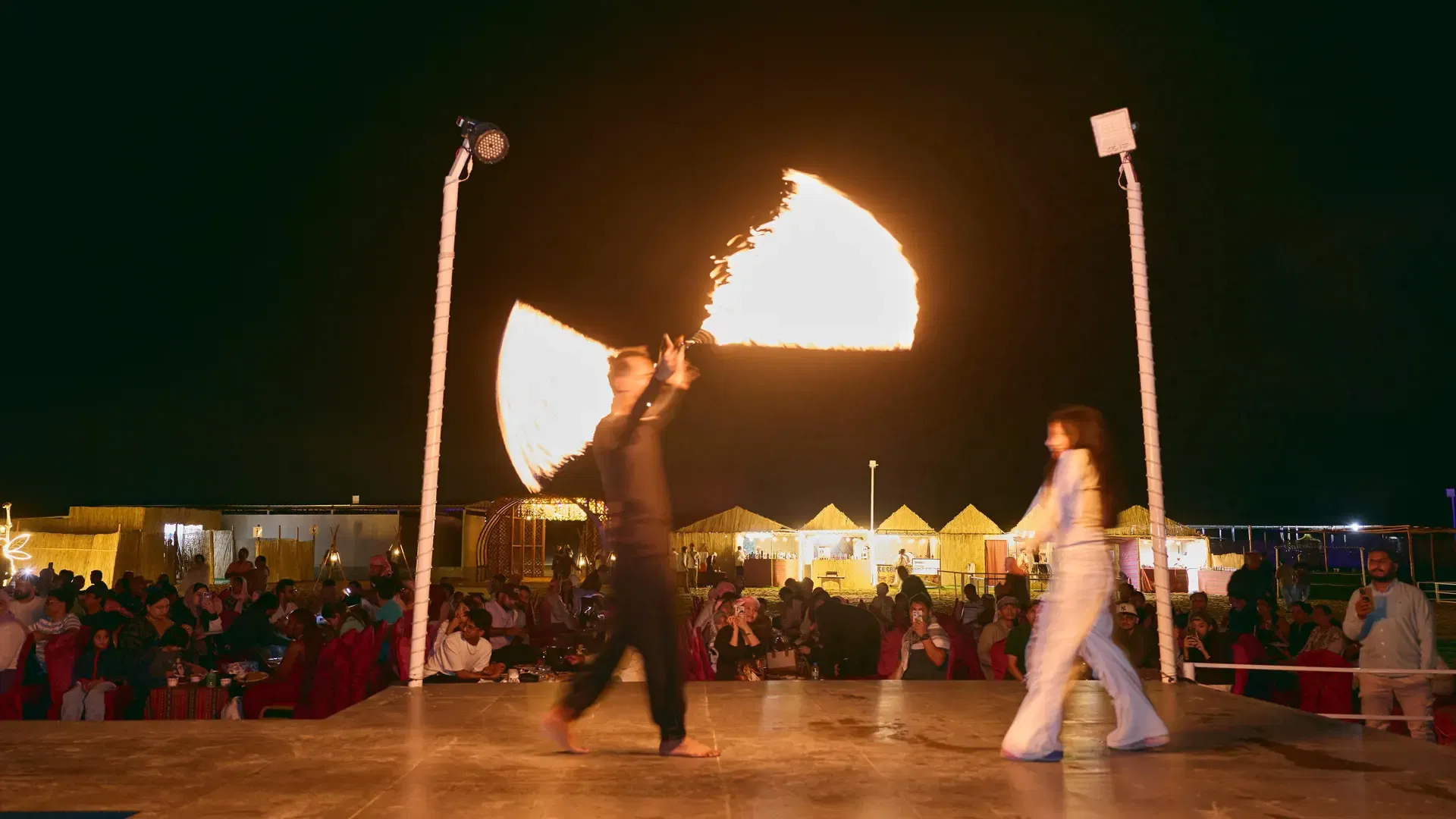 A male fire performer holding two burning flails and a female performer in white walk across an illuminated stage at night at Desert Safari + Al Faqaa Camp.