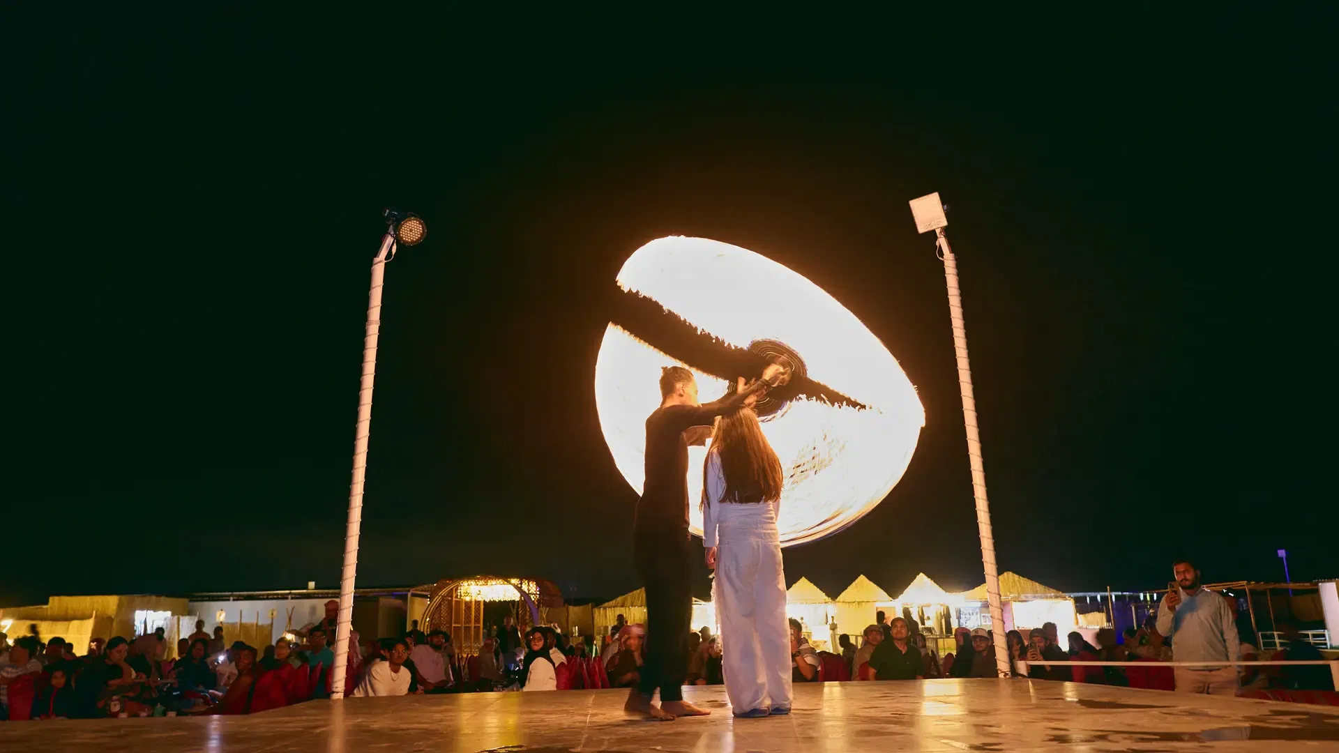 Two performers stand on stage with a large glowing circular fire effect spinning behind them at night at Desert Safari + Al Faqaa Camp.