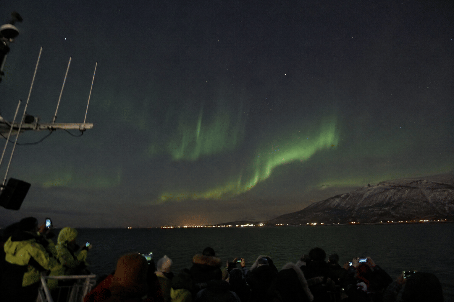 Faint green Northern Lights stretching over distant mountains and city lights at night.