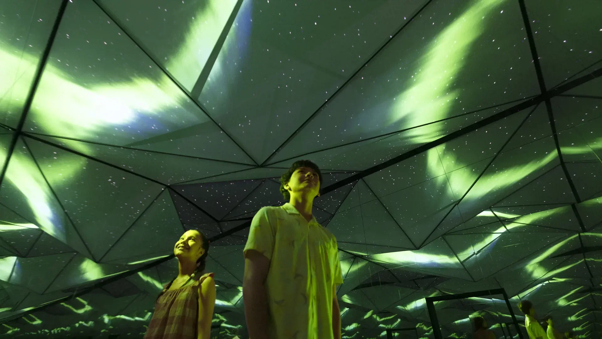 Two people stand looking upward at a faceted ceiling lit with green glowing patterns resembling northern lights at Exploria in Mandai Singapore Ticket.