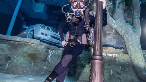 A scuba diver in full gear leans against a decorative underwater lamppost with a submerged car parked on a ledge behind them at Deep Dive Dubai.