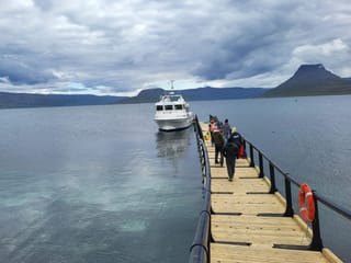 A jetty leading into the forground where a boat sits alongside and a mountain view in the background