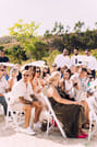 A wide shot of wedding guests seated in rows of chairs, looking toward the back of the venue in anticipation. The ceremony space is decorate