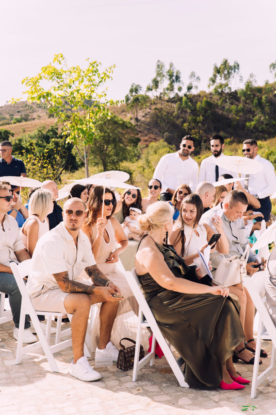 A wide shot of wedding guests seated in rows of chairs, looking toward the back of the venue in anticipation. The ceremony space is decorate