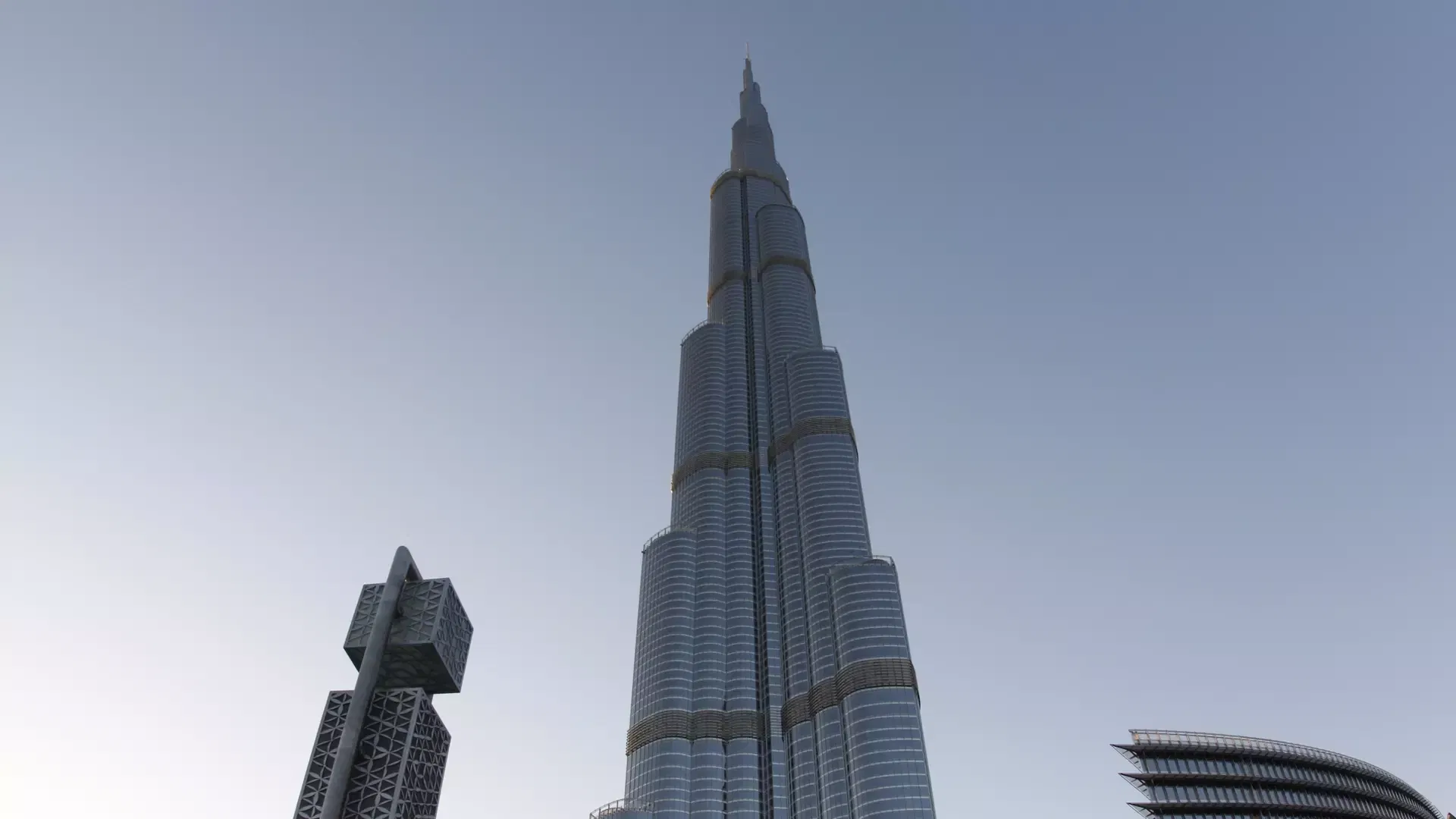The full tapering height of the tower against a soft blue evening sky, with a lattice-clad sculpture and a curved glass building in the foreground at Burj Khalifa
