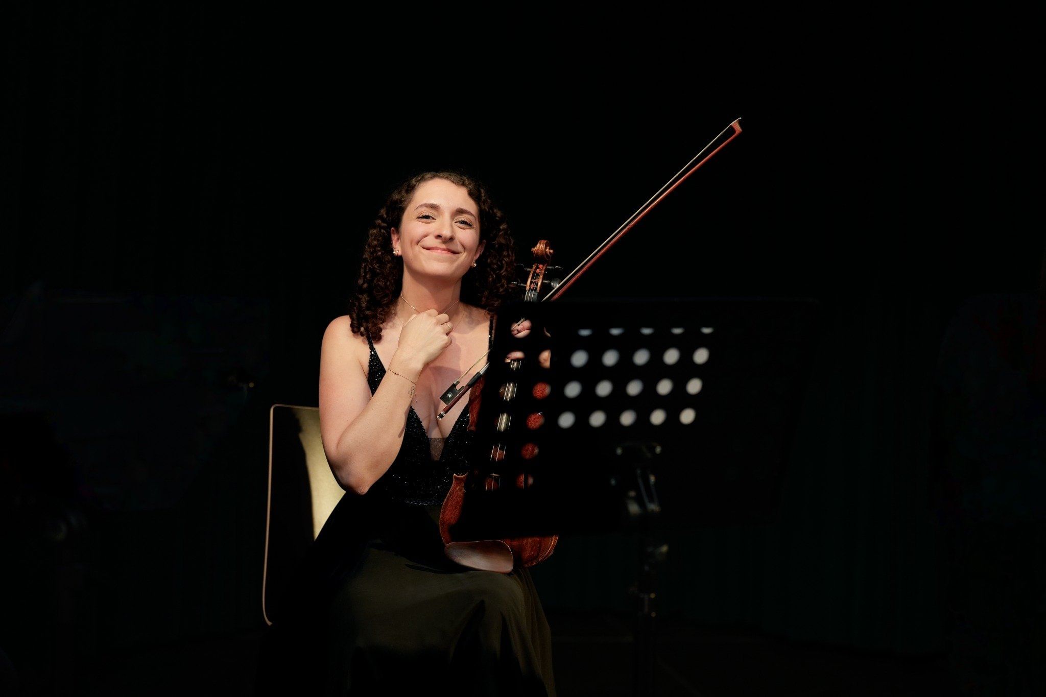 Woman seated with a violin and bow, smiling under stage lighting.