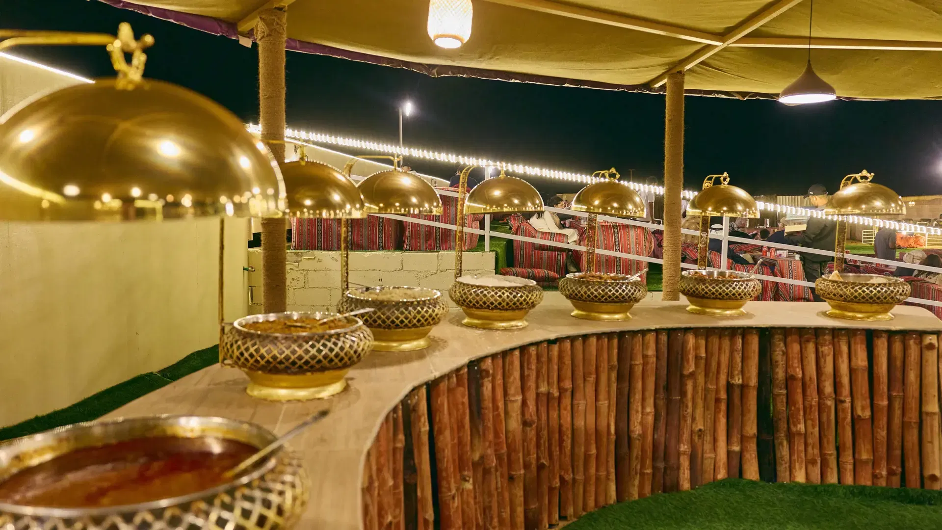 A buffet counter features multiple golden serving bowls under a canopy tent at night at Desert Safari + Al Faqaa Camp.