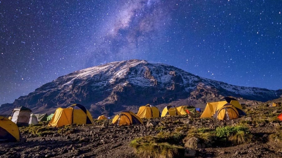 Yellow expedition tents illuminated at night in a high-mountain camp with Mount Kilimanjaro and the Milky Way overhead.
