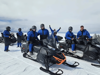 Group of eight visitors in matching blue snowmobile suits and helmets posing with Arctic Cat snowmobiles on the flat white surface of Langjo