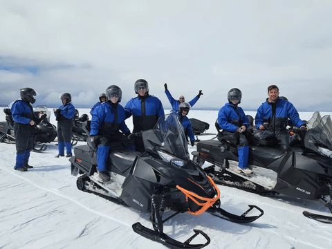 Group of eight visitors in matching blue snowmobile suits and helmets posing with Arctic Cat snowmobiles on the flat white surface of Langjo